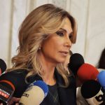 Femeie blondă răspunde presei, înconjurată de microfoane.
